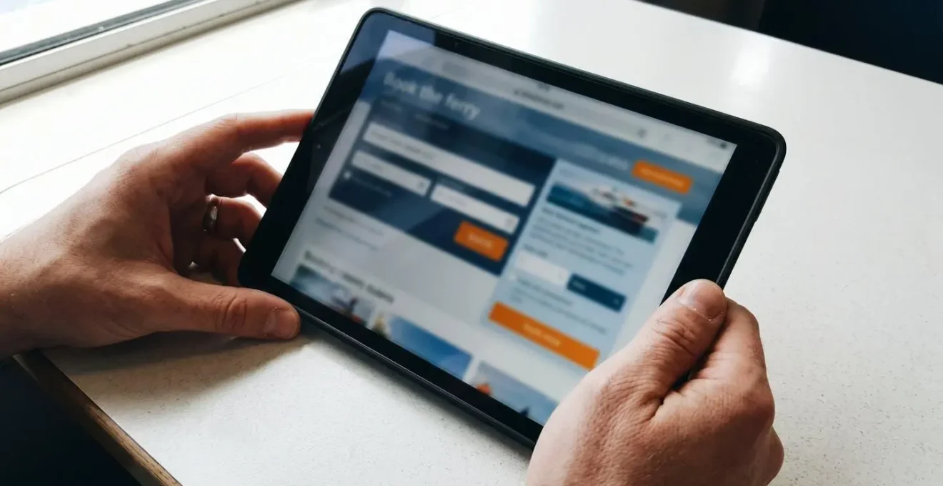 Mani di una persona che consulta un tablet mostrando uno schermo sfocato di prenotazione online, su un tavolo moderno con luce naturale